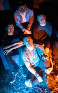 The four men who are members of The Engagement sit on the ground looking up at the camera which is above them